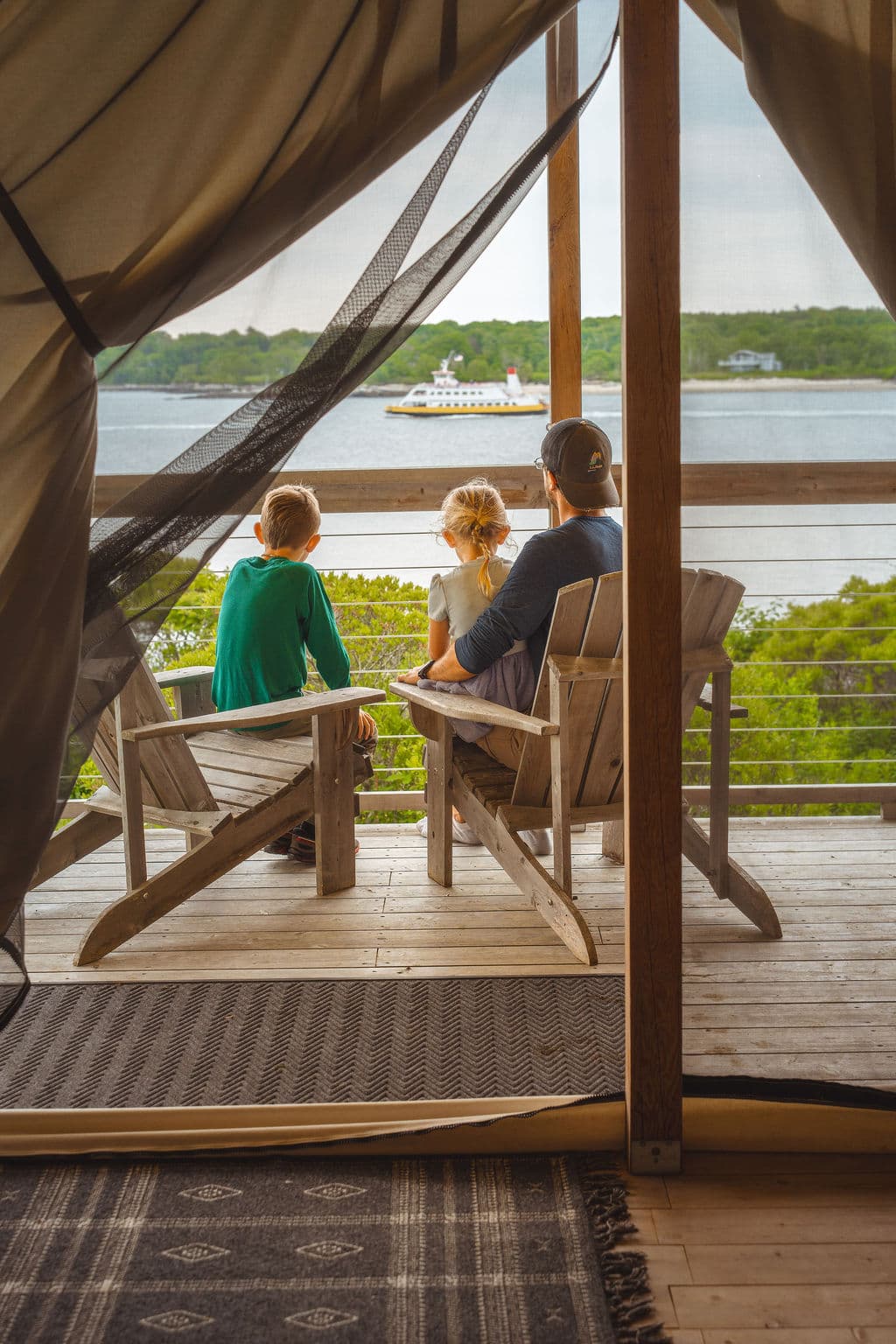 A father and two children sit on a porch overlooking a serene water view.