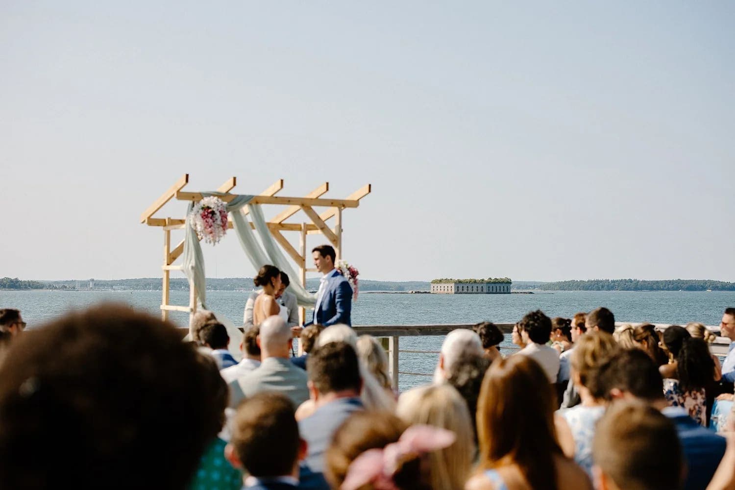A couple exchanges vows under a floral-adorned arch at a lakeside wedding ceremony with guests seated nearby.