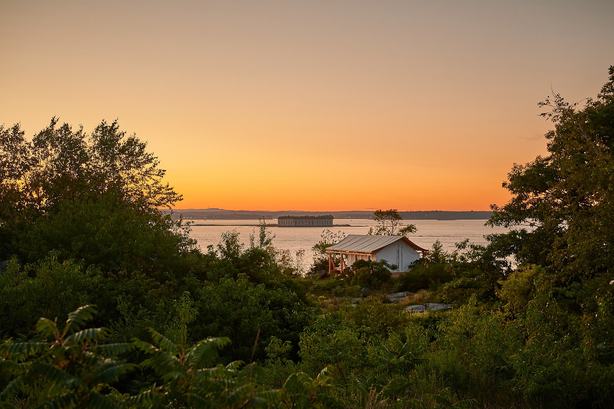 A serene landscape at sunset, featuring a house surrounded by trees with a calm water view.