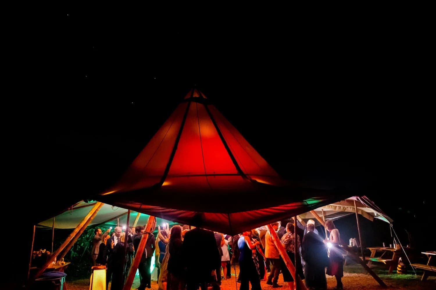 A vibrant gathering under a red-tented structure at night, surrounded by softly glowing lights.