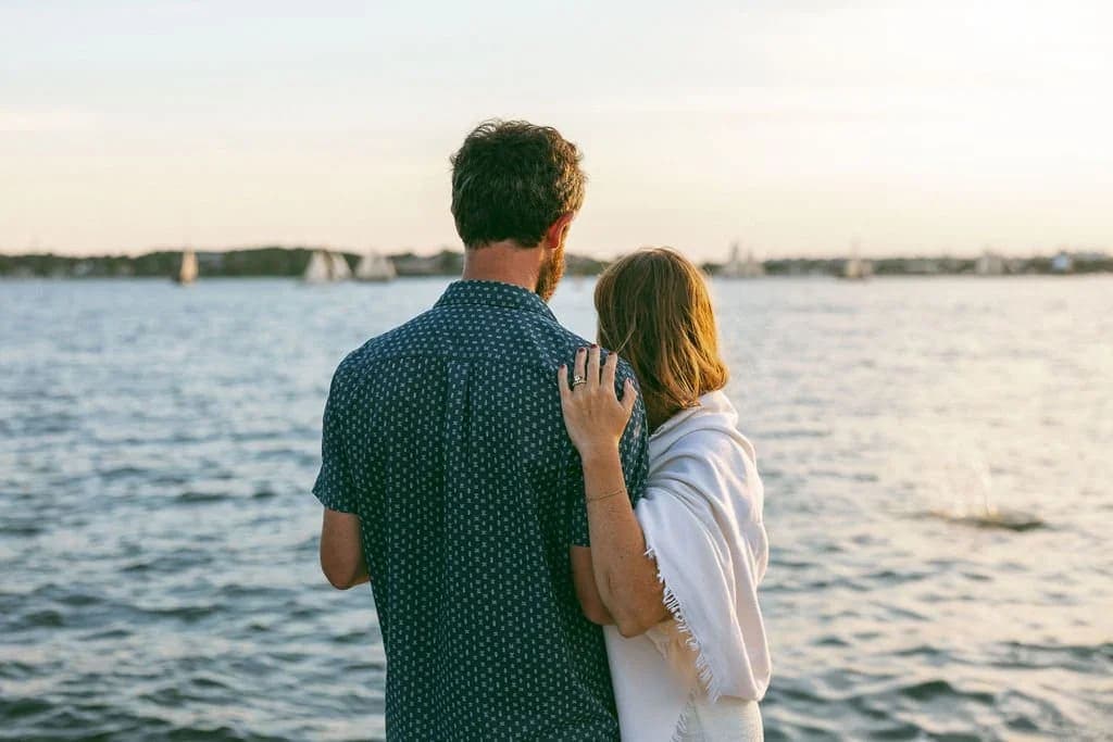 A couple stands by the waterfront, gazing at the sunset over the water.