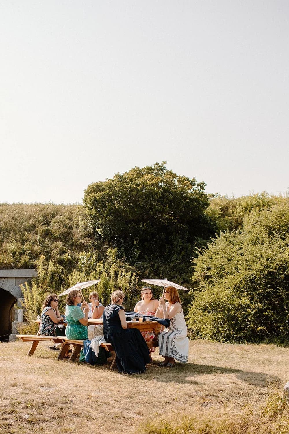 A group of six women sitting at a picnic table under parasols in a grassy outdoor setting.