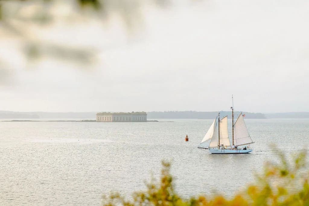 A sailing boat with white sails glides across a calm sea near an island with a historic building.