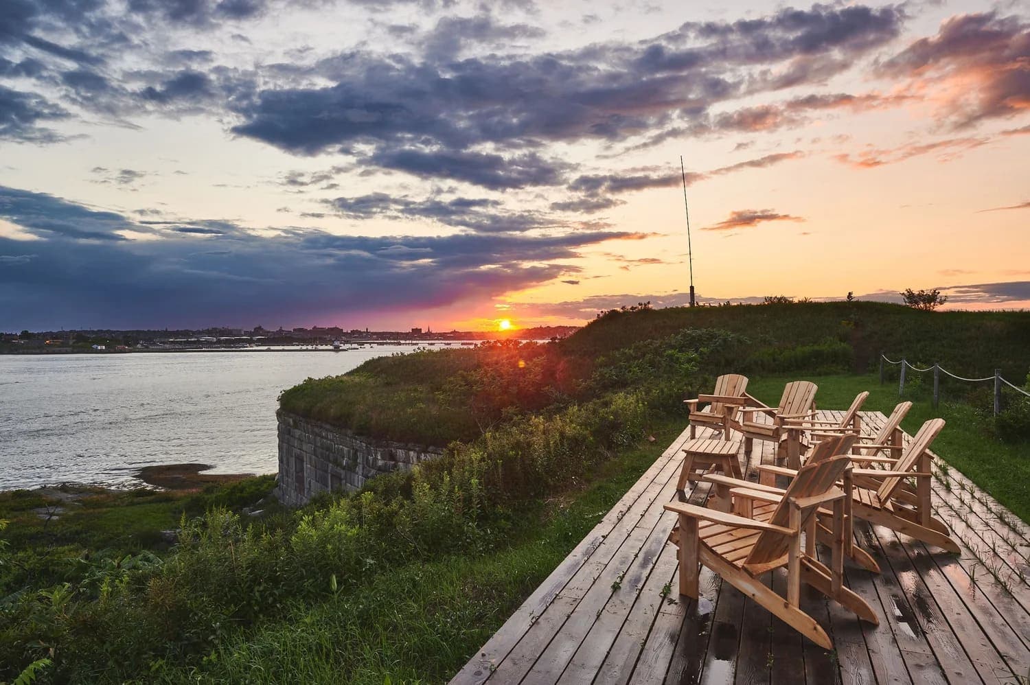 Adirondack chairs overlook a river at sunset, with dramatic clouds in the sky.