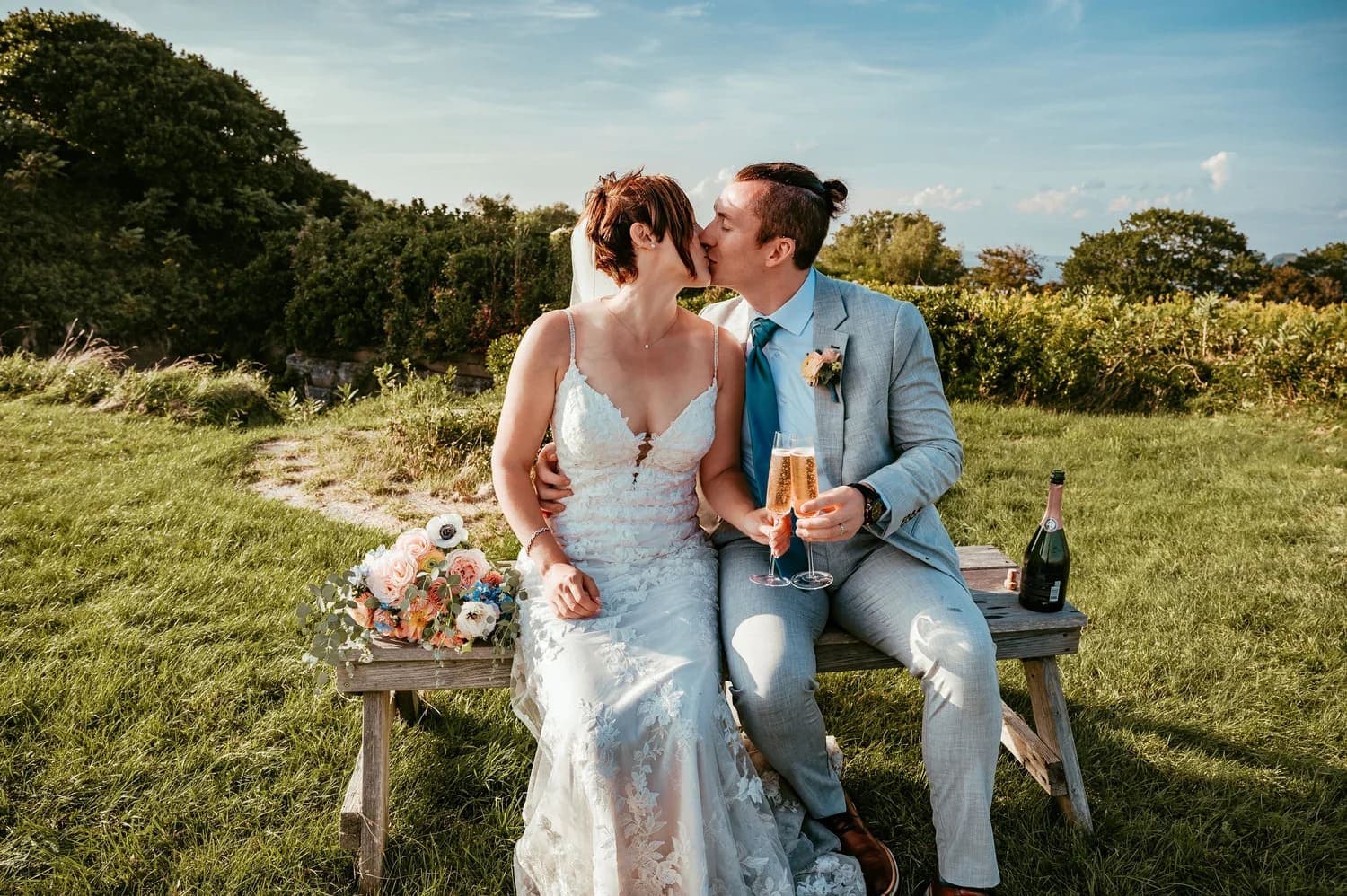 A couple in wedding attire sharing a kiss while holding champagne glasses, seated on a bench in a scenic outdoor setting.