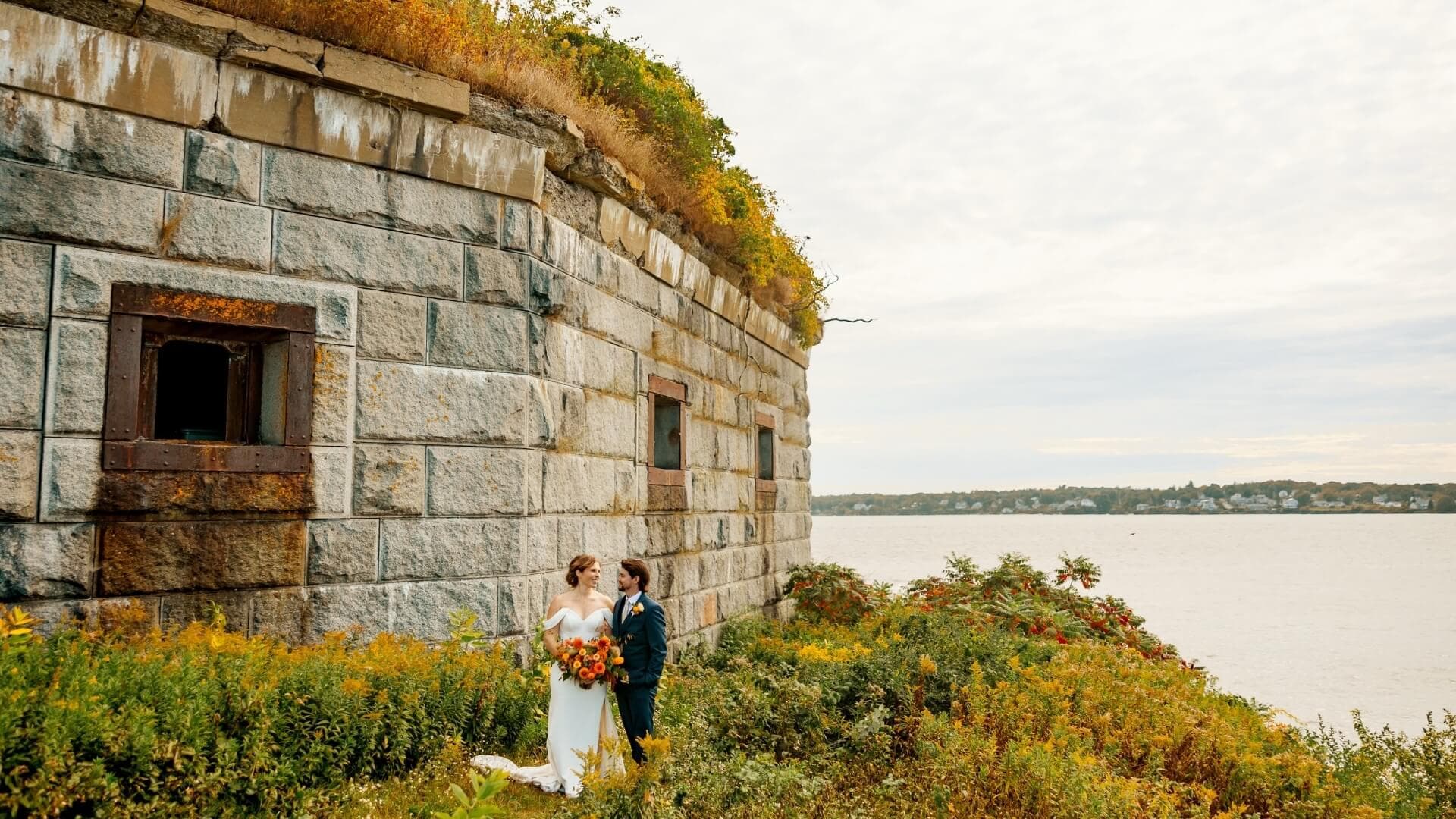 A couple in wedding attire stands together by an old stone fort overlooking a calm water body.