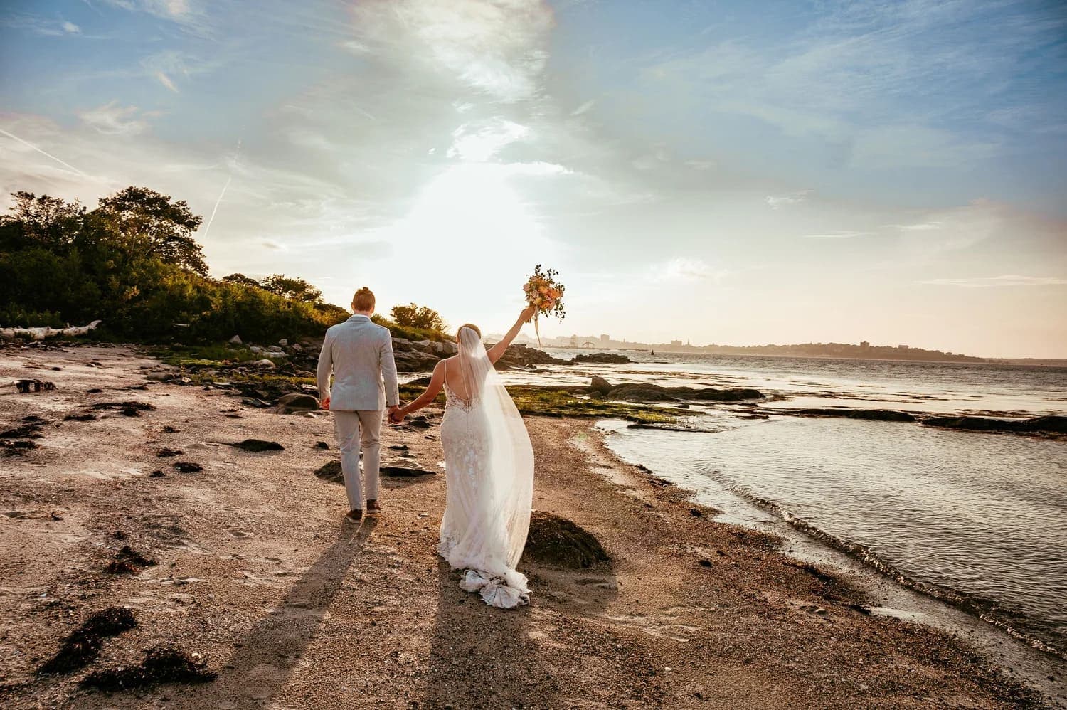 A newlywed couple walks hand-in-hand along a beach at sunset.