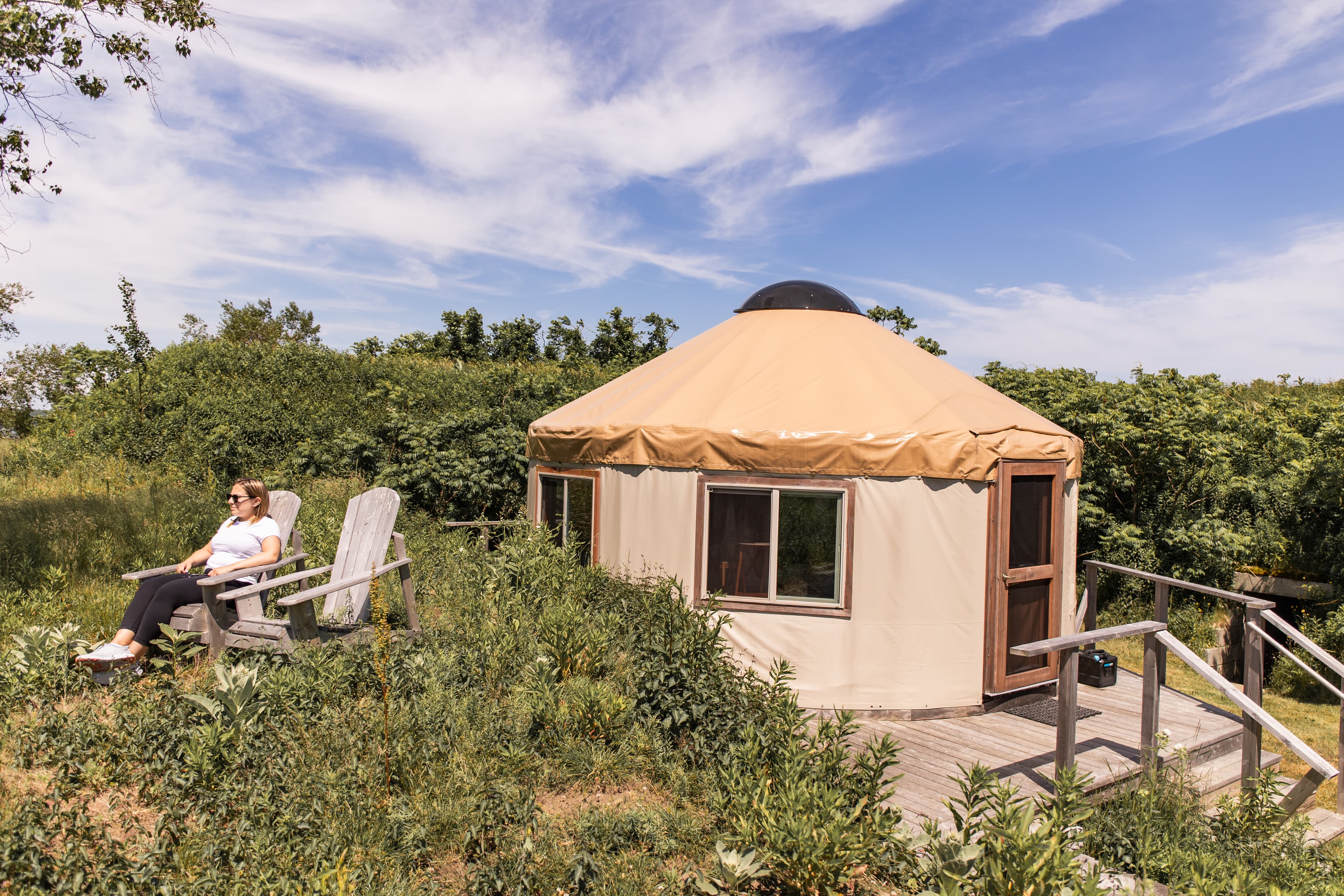 A person relaxes in an outdoor chair near a yurt surrounded by greenery under a blue sky.