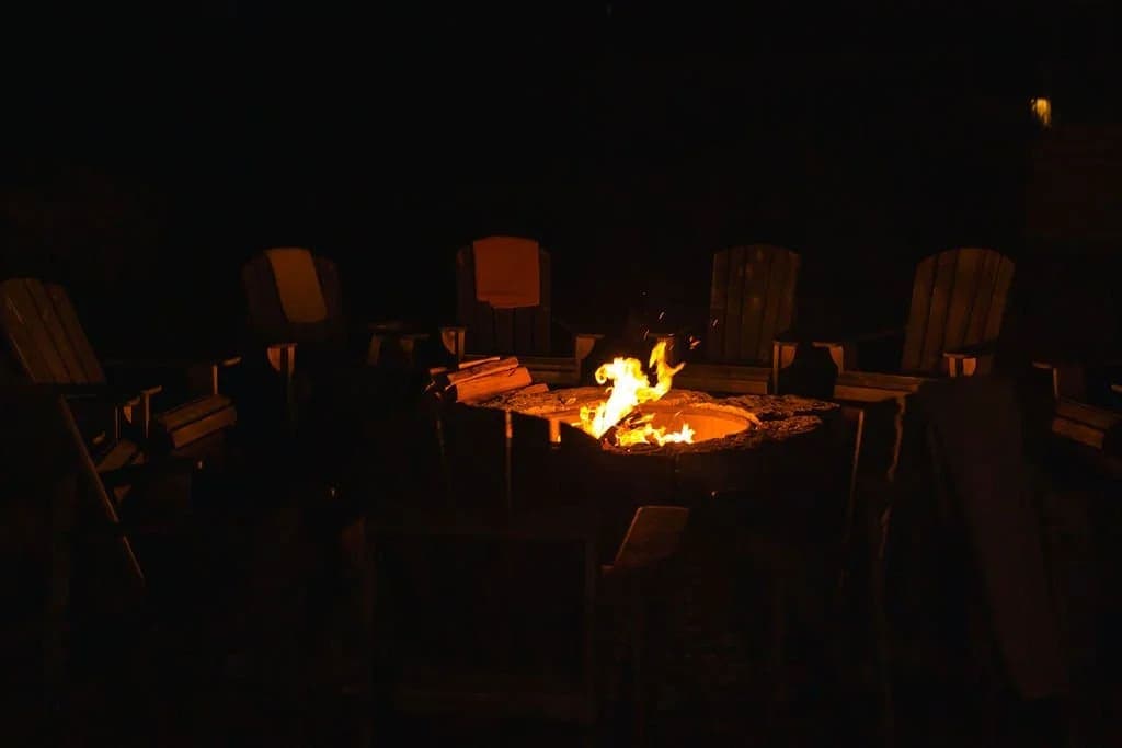 A warm fire burns in a stone pit surrounded by wooden chairs at night.