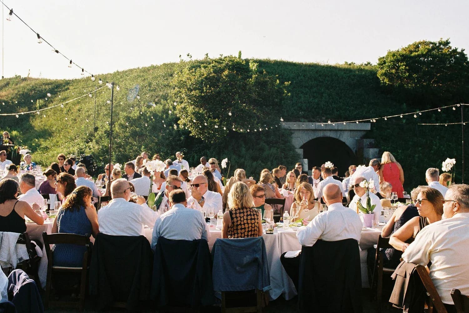 A large outdoor gathering with guests seated at tables adorned with flowers, enjoying a meal under string lights.