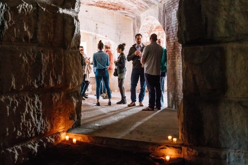 A group of people stands in a dimly lit space with stone walls, gathered around candles.