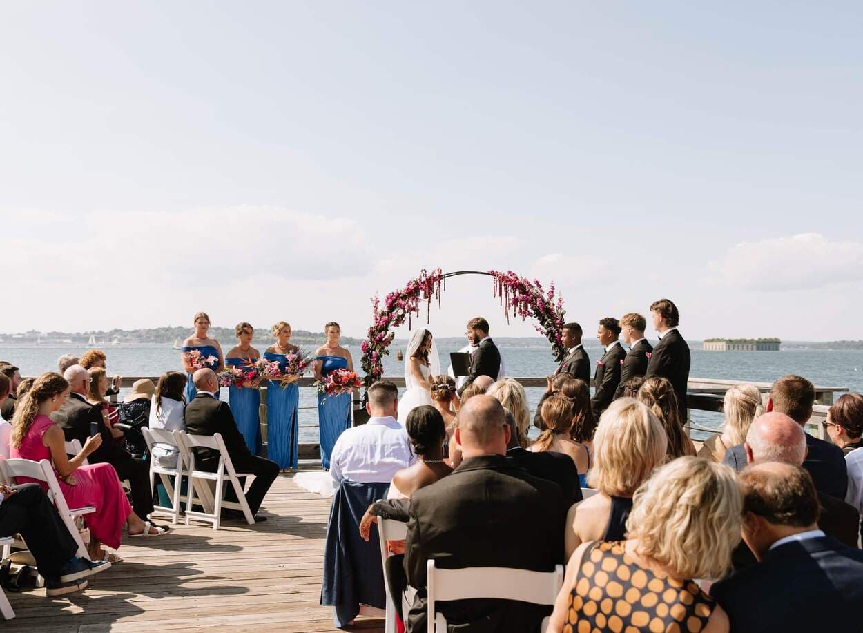 A wedding ceremony takes place outdoors by the water, surrounded by guests and a wedding party.