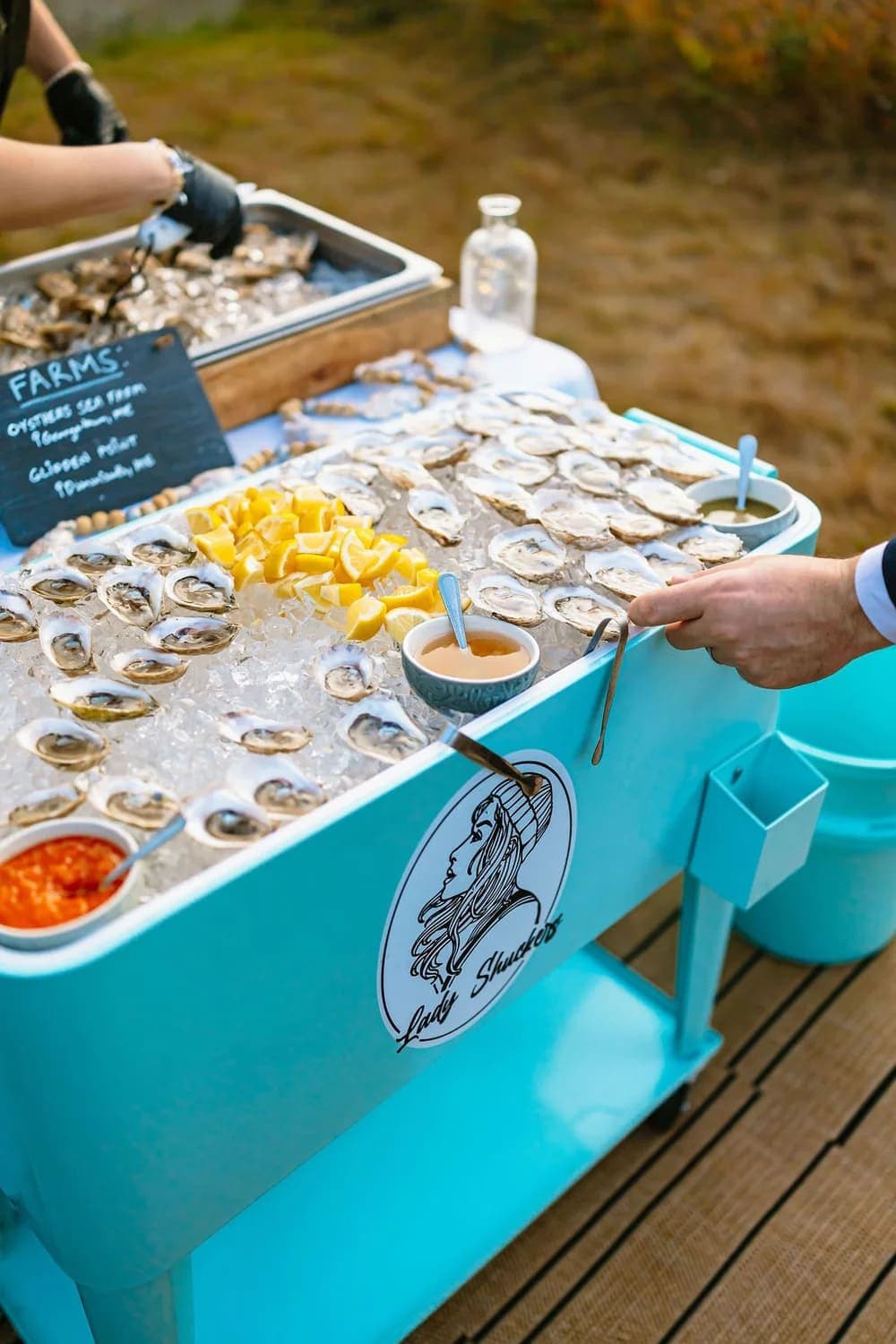 A hand reaches for an oyster on ice at a seafood stand display featuring lemons and sauces.
