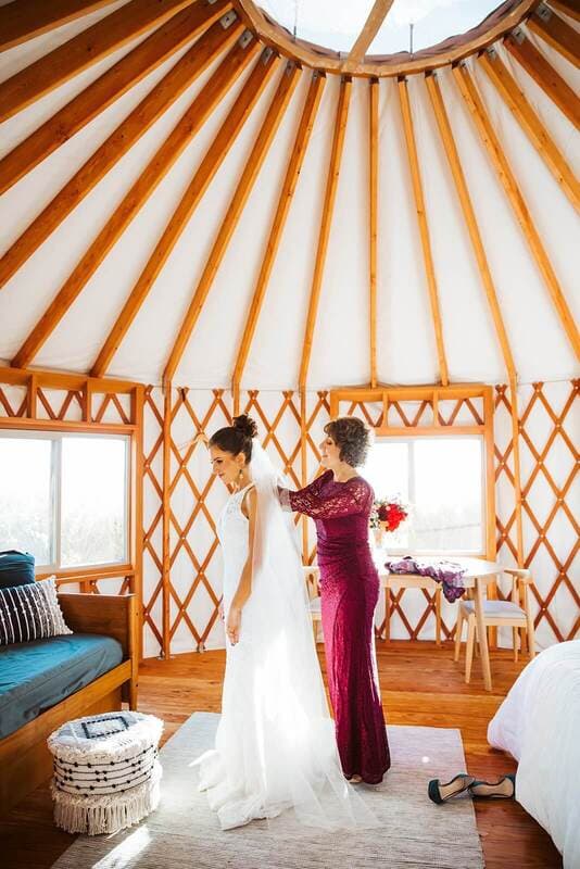 A bride is adjusting her veil with the help of a friend in a cozy, wooden yurt.