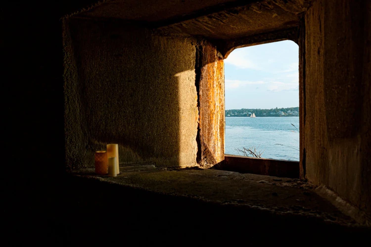 Light streams through a rectangular window with candles, overlooking a serene body of water and distant sailboat.