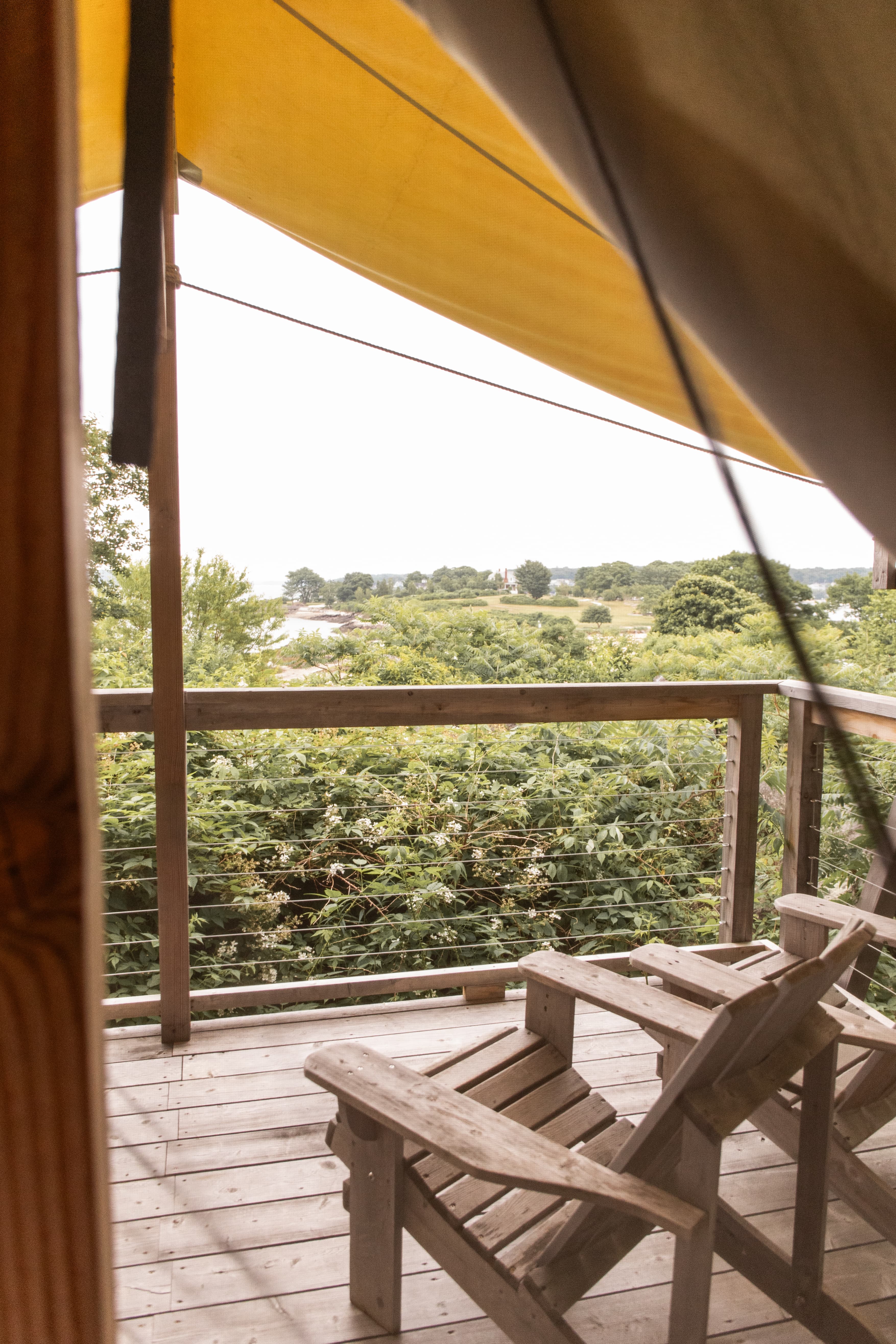A wooden balcony with two chairs overlooks a lush green landscape under a yellow canopy.