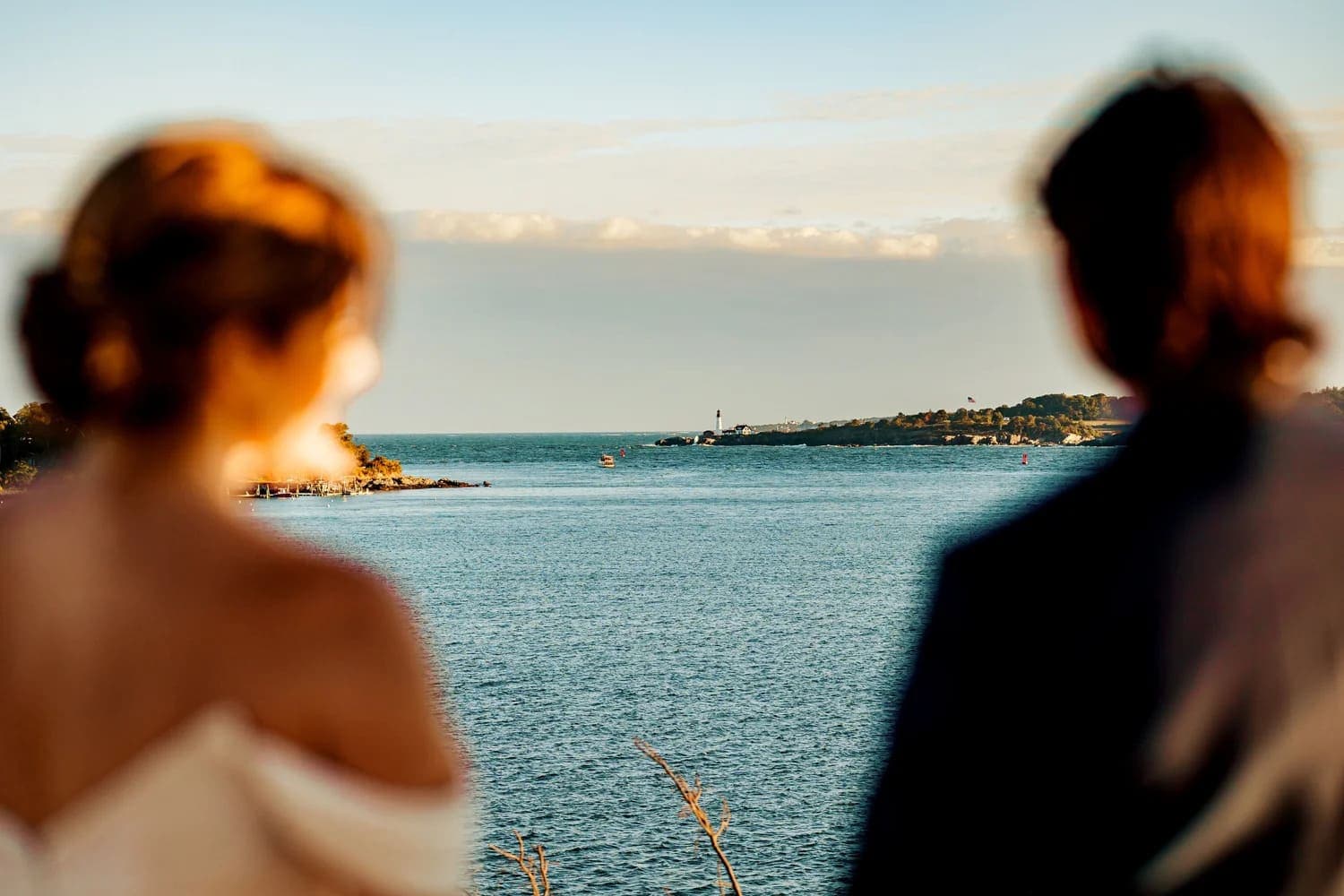 A couple stands together, gazing at a scenic waterfront view with a lighthouse in the distance.