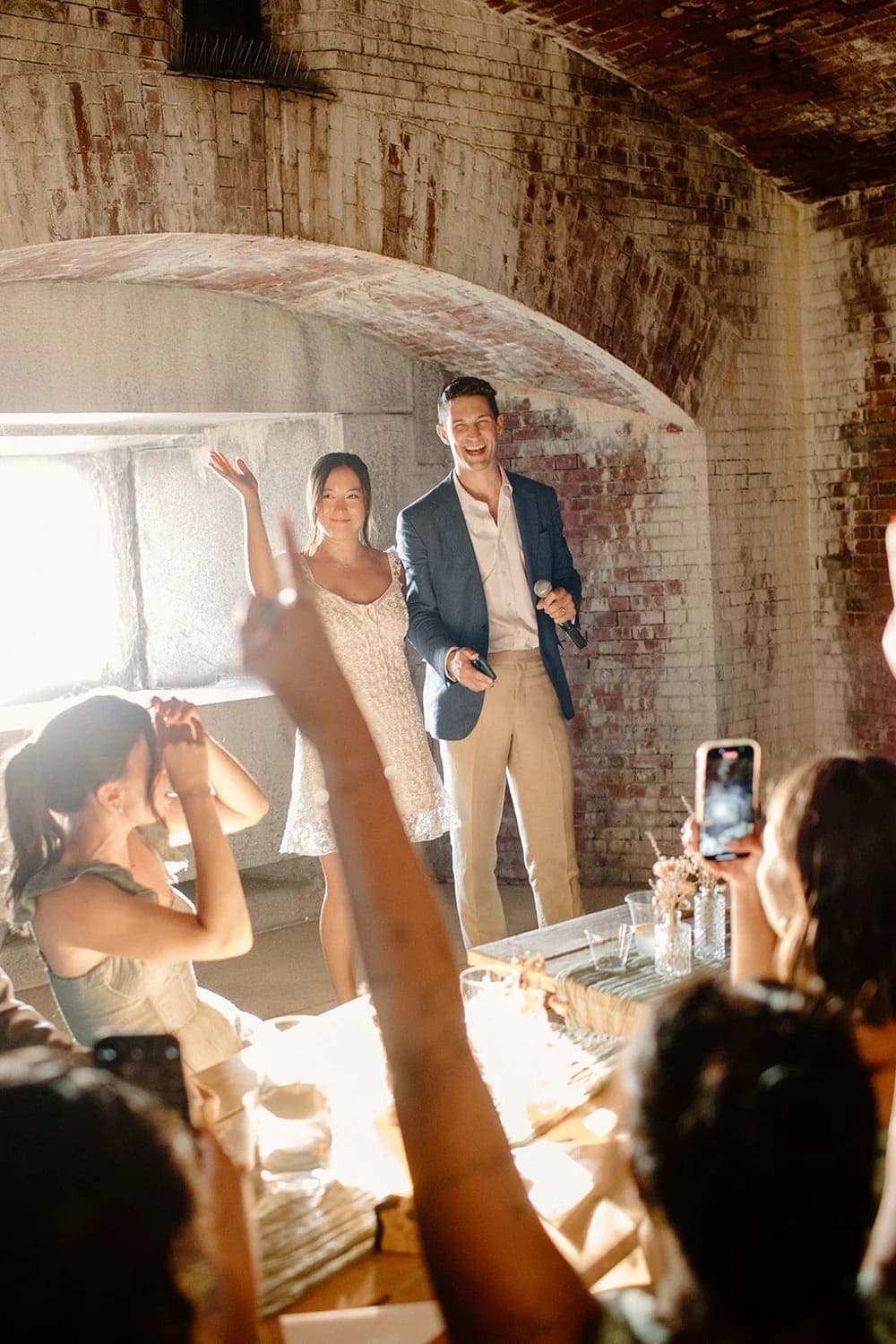 A couple joyfully greets friends in a rustic setting, as guests celebrate with raised hands and phones.