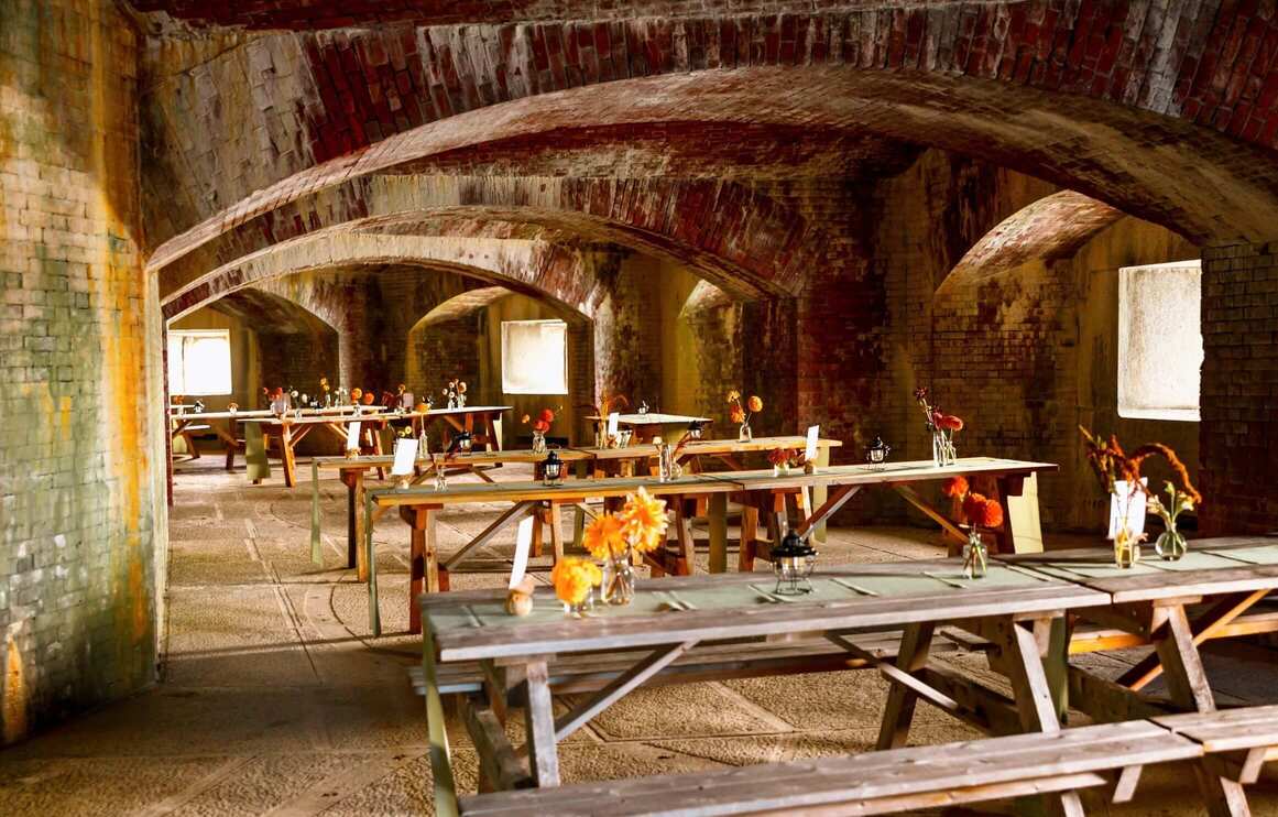 A rustic dining space with wooden tables decorated with flowers under arched brick ceilings.