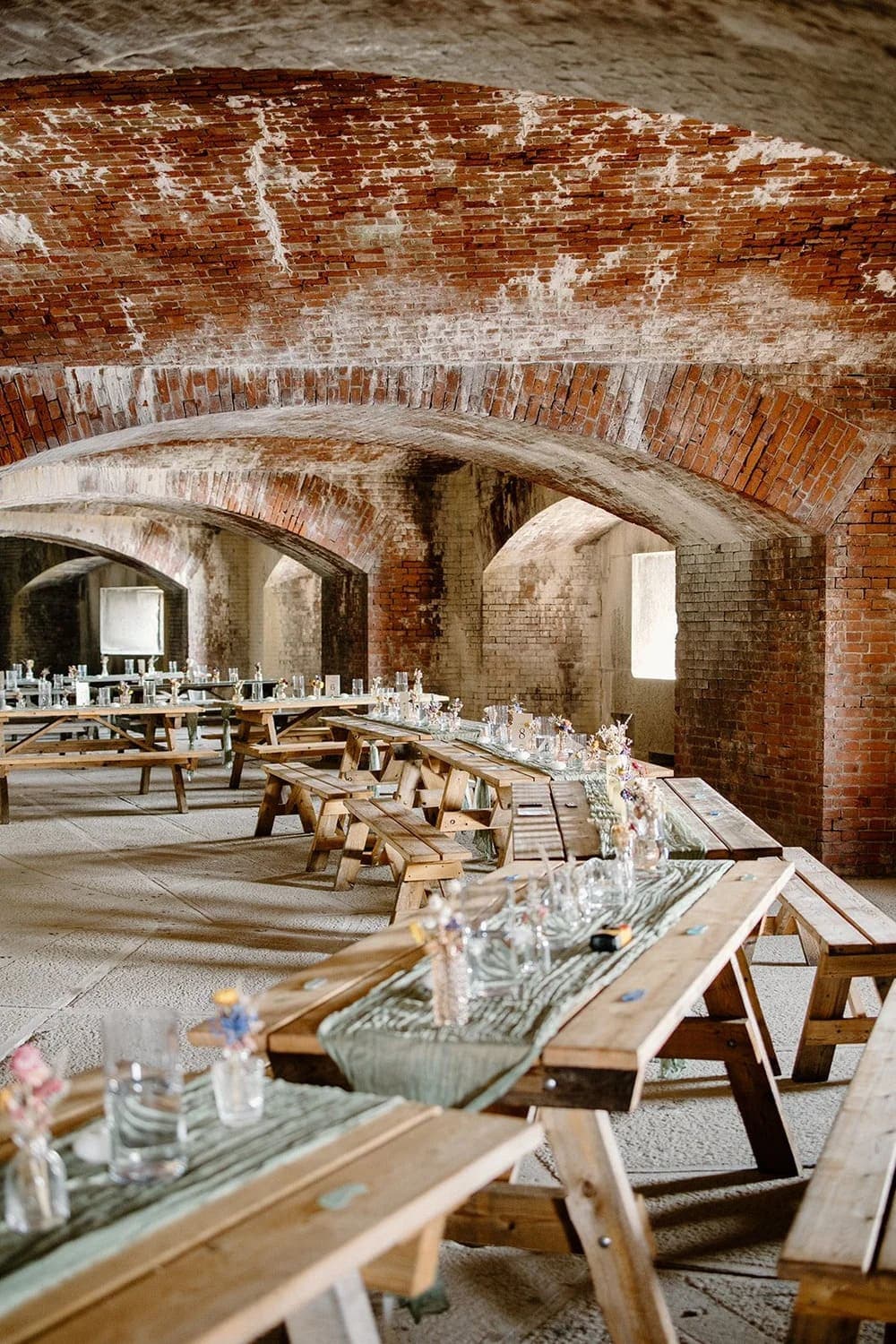 A rustic dining area with wooden benches and tables adorned with glassware, set under a brick archway.