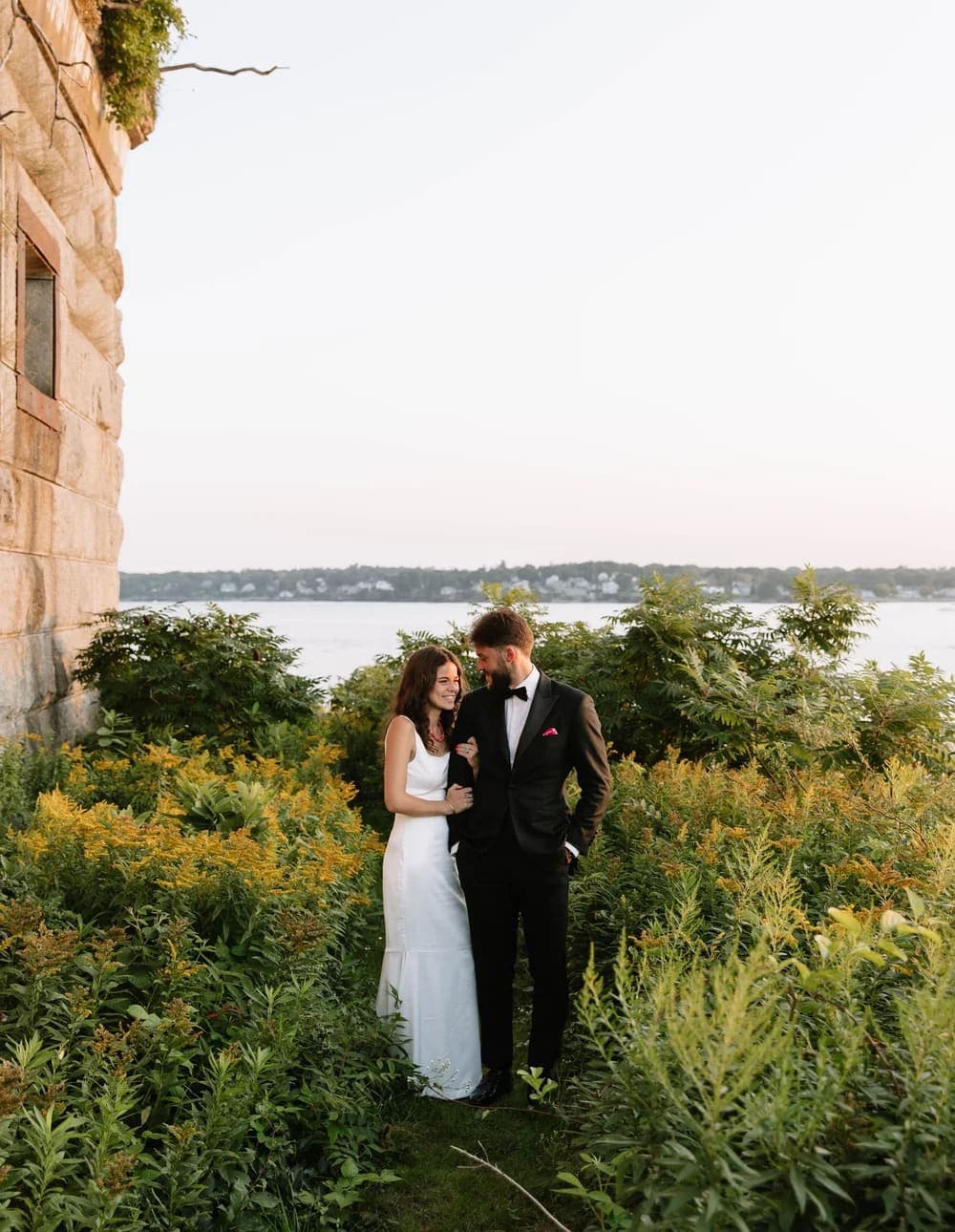 A couple embraces in a lush green area near a serene body of water at sunset.