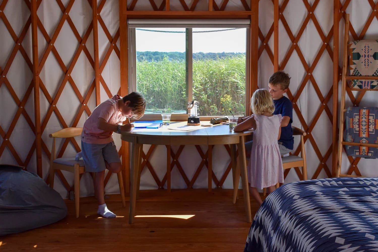 Two children are engrossed in activities at a round table with a window view of green fields.