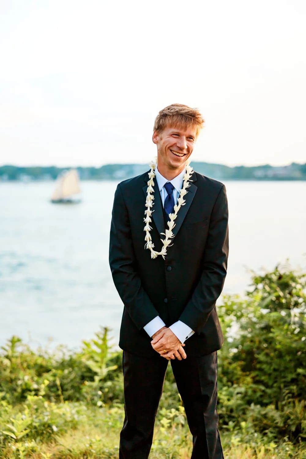 A smiling man in a suit with a floral lei stands by the water.