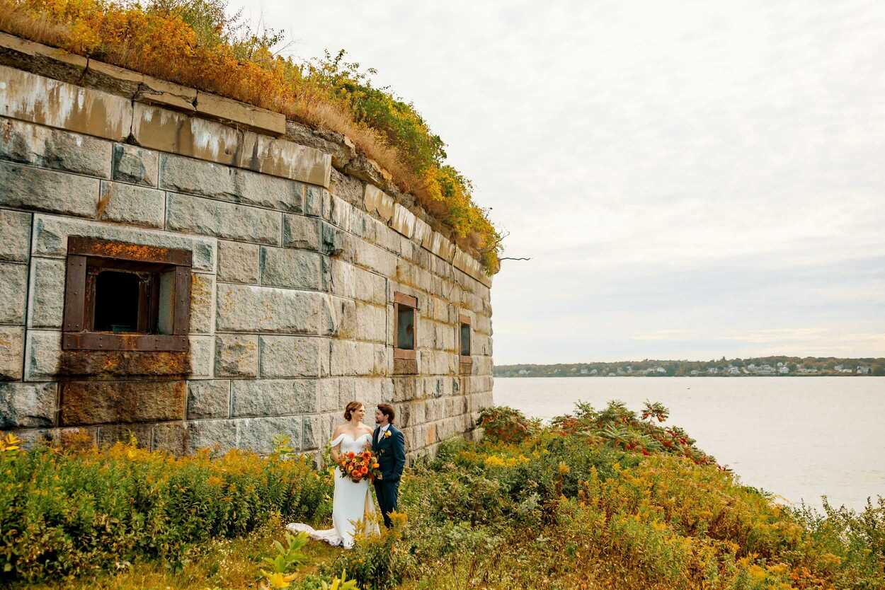 A bride and groom stand together by a stone wall overlooking the water.