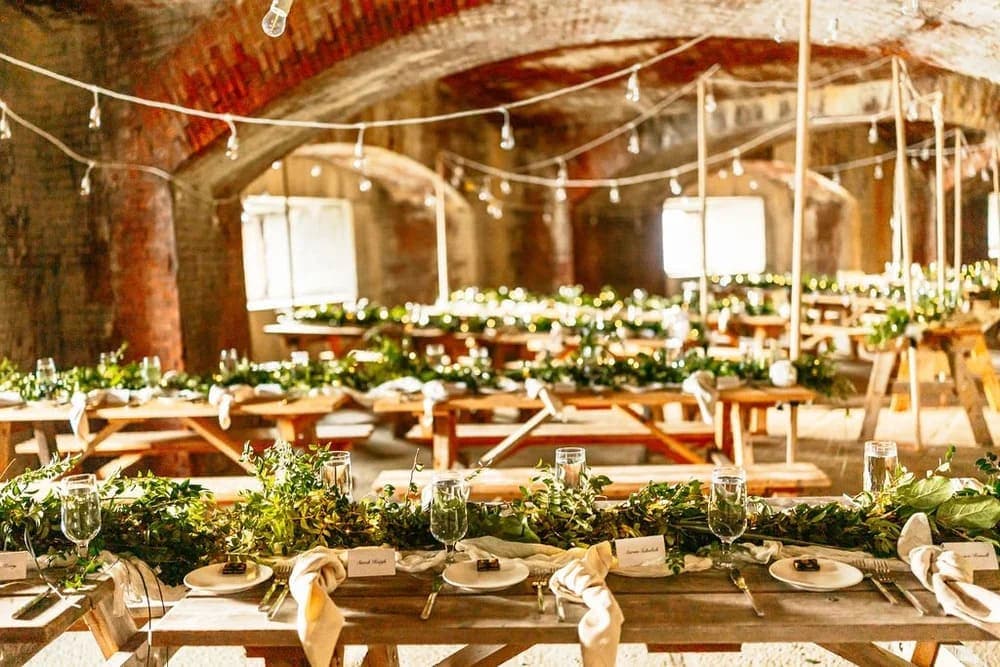 A beautifully arranged banquet hall with wooden tables adorned with greenery and elegant place settings, illuminated by hanging lights.