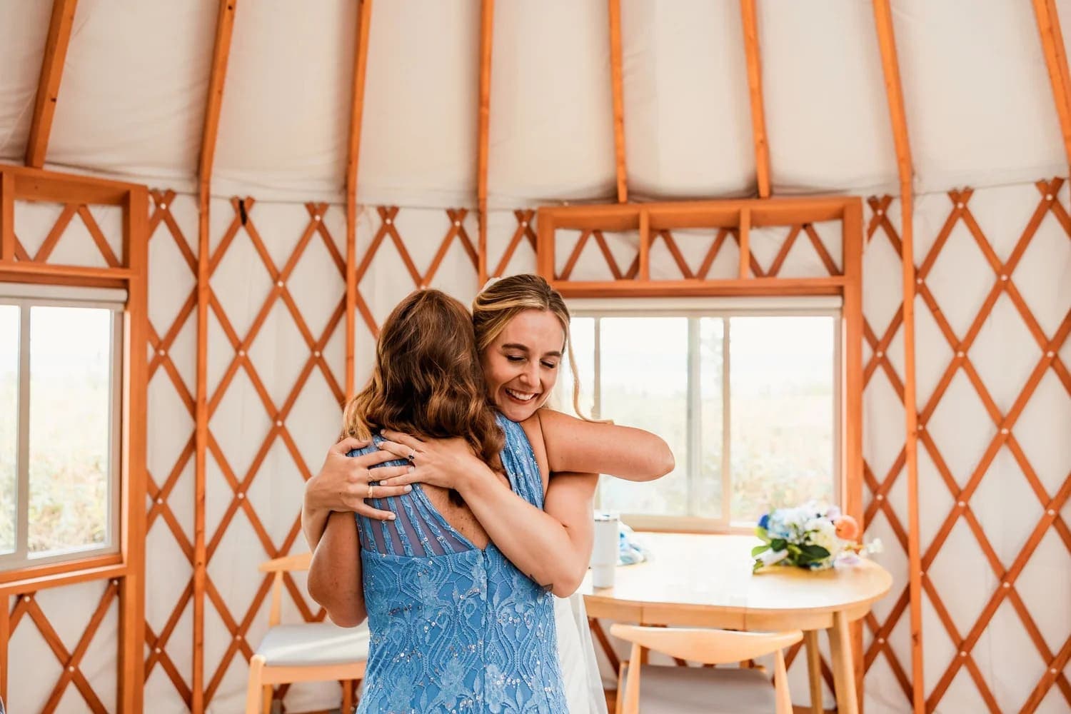 Two women happily embrace inside a yurt, with a round table and floral arrangement in the background.