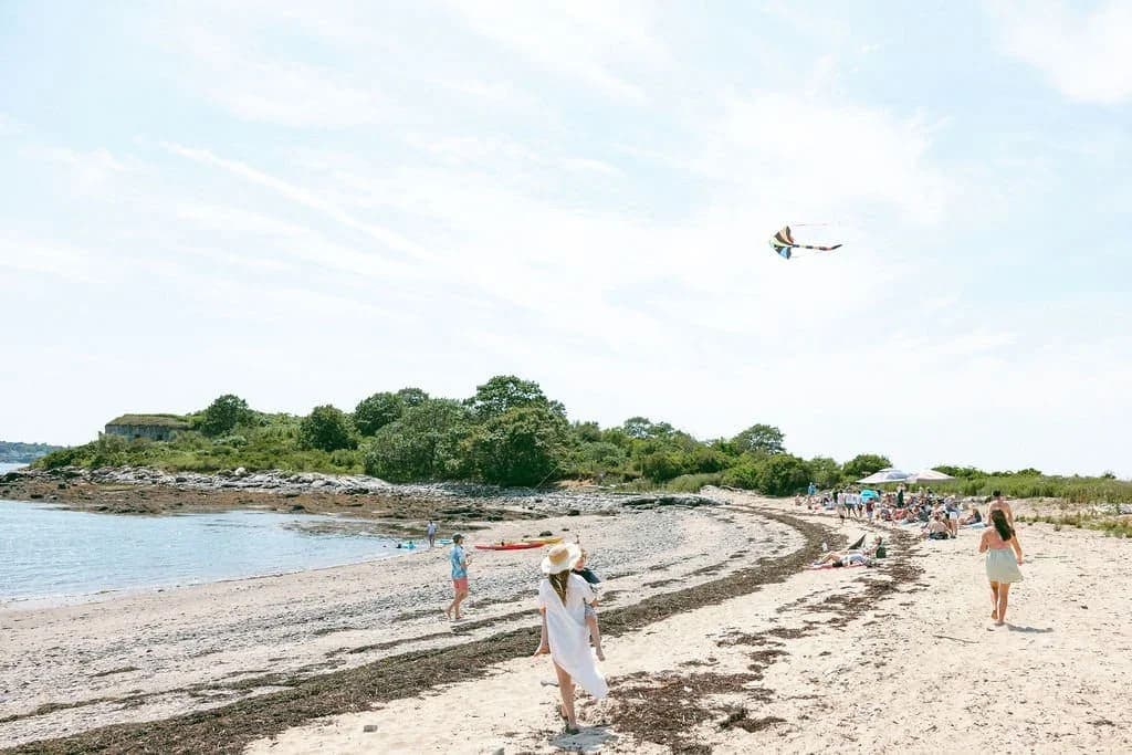 People enjoying a sunny day on a sandy beach, with a kite flying overhead.