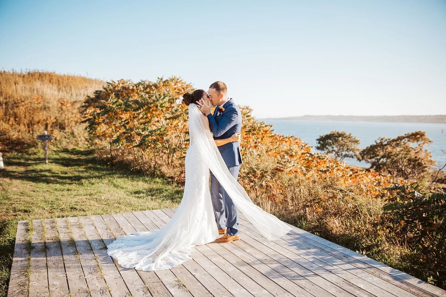 A newlywed couple shares a kiss on a wooden platform overlooking a scenic landscape.