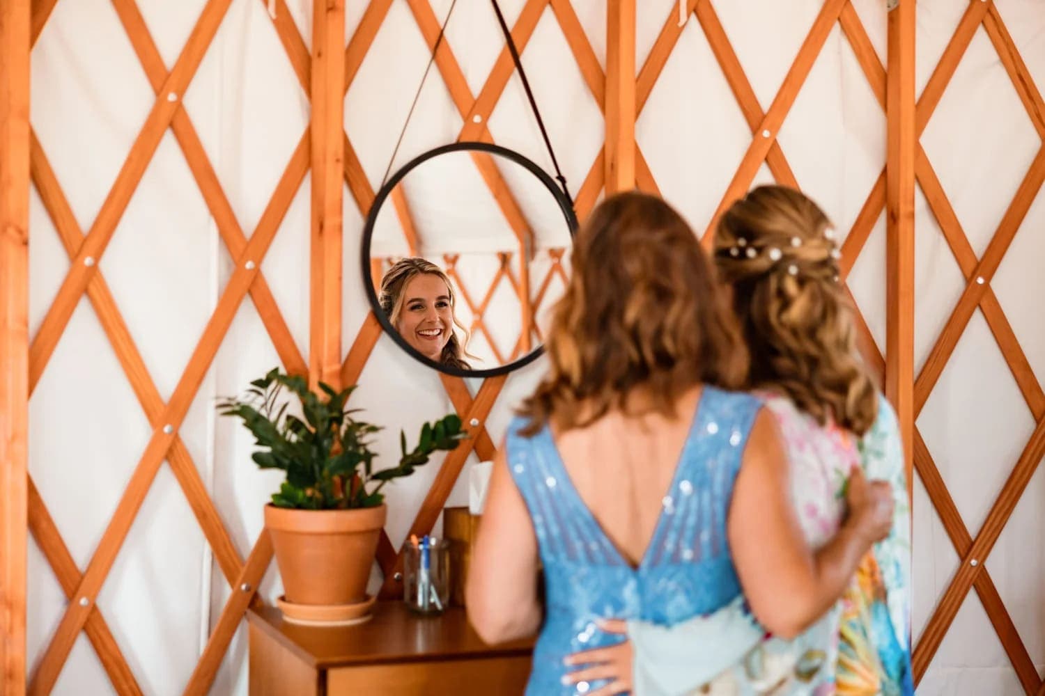 A woman smiles at her reflection in a mirror while another woman stands beside her, both in a warmly decorated setting.