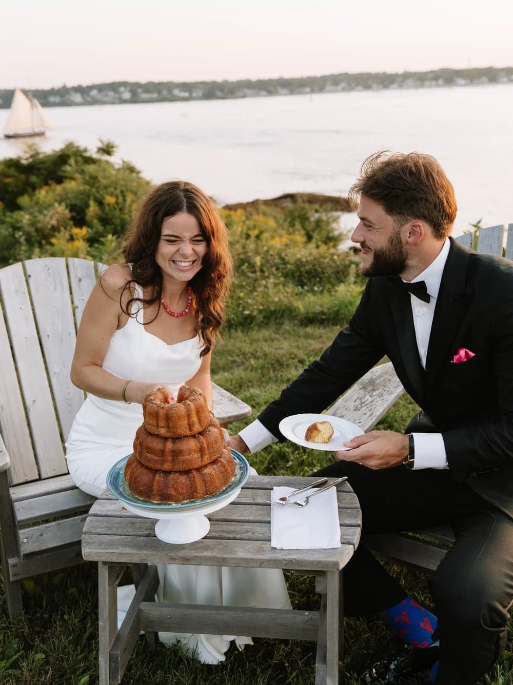 A joyful bride and groom share a dessert during sunset by the water.