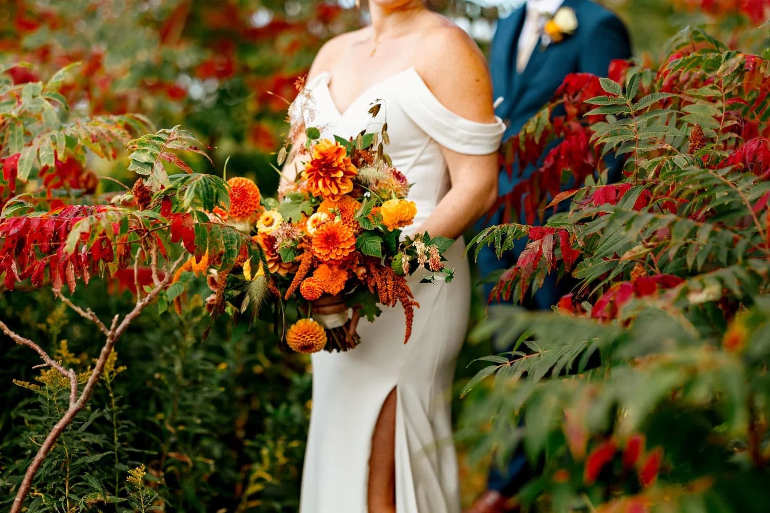 A bride in a white gown holds a vibrant bouquet among colorful autumn foliage.