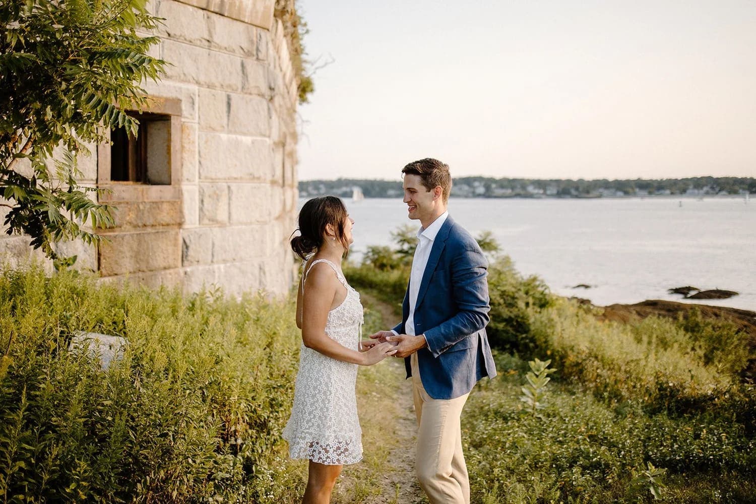 A couple stands holding hands by the water, surrounded by greenery and an old stone structure.