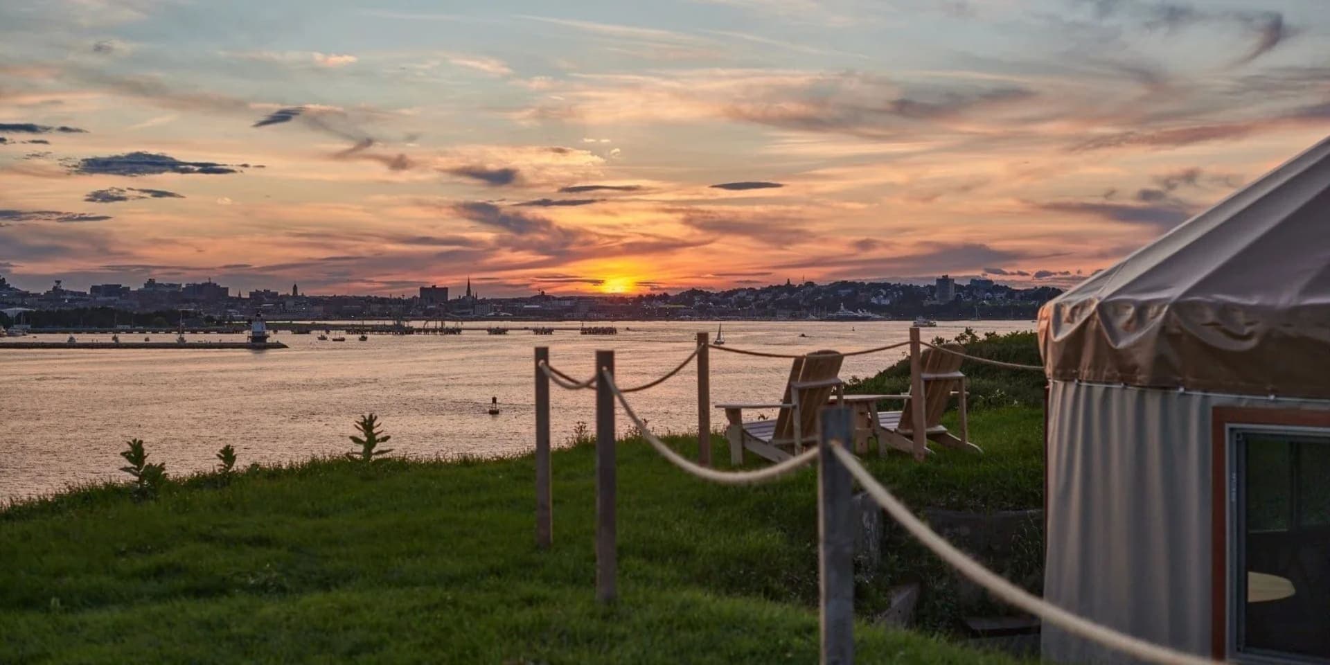 A serene sunset over a river, with a grassy shoreline and a tent in the foreground.