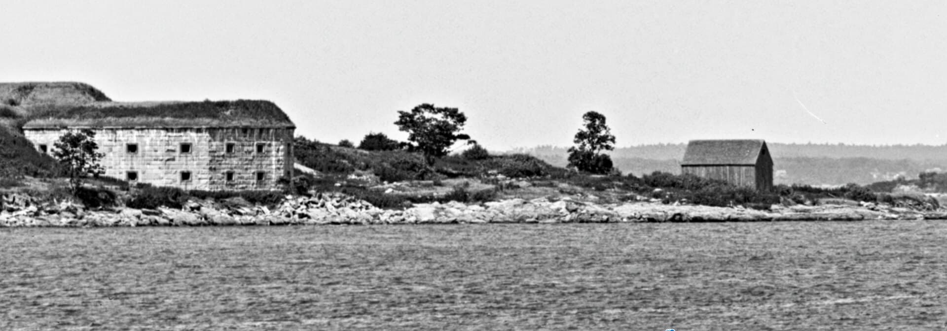 Black-and-white photo of two weathered buildings on a rocky shoreline with sparse vegetation.