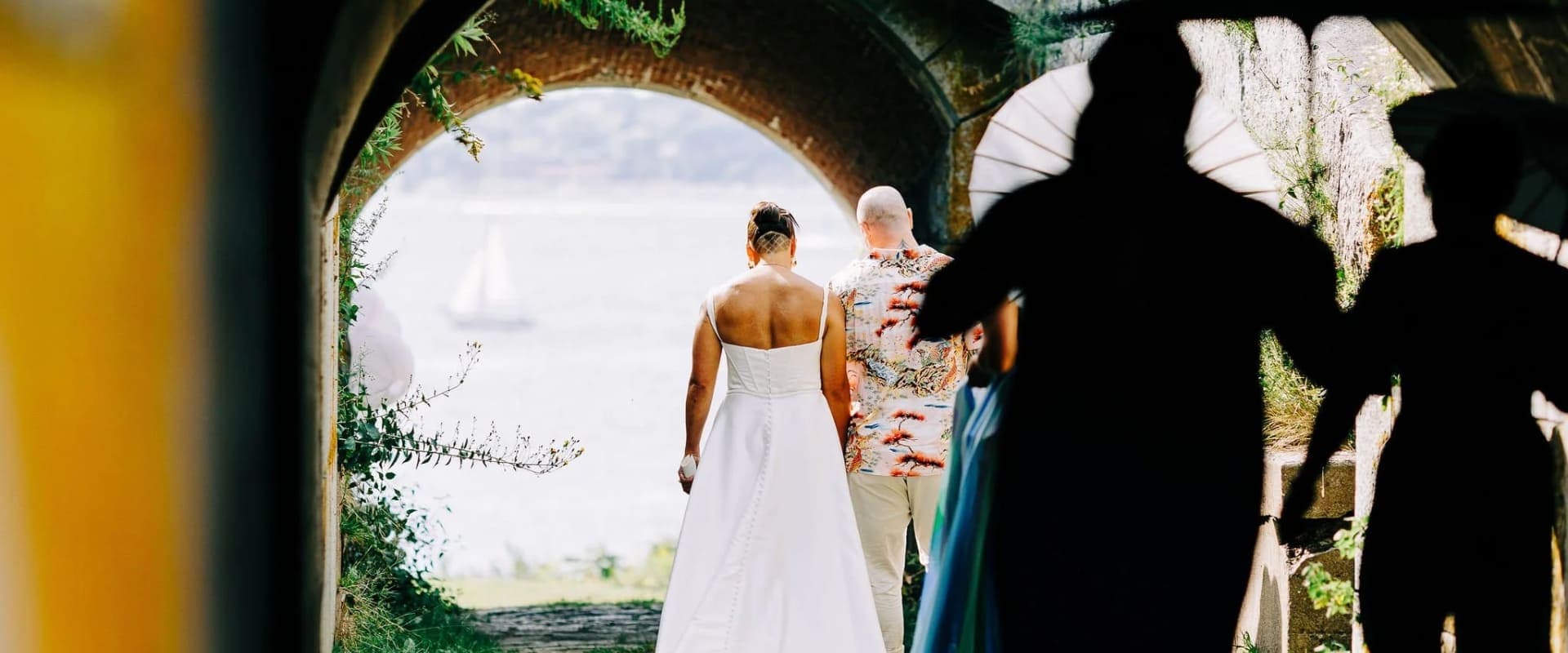 A bride and groom stand together under an archway, with silhouettes of guests in the foreground and a scenic view in the background.