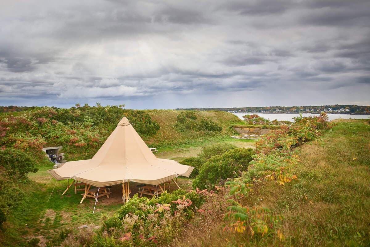 A large canvas tent is set on a grassy area with water and overcast skies in the background.