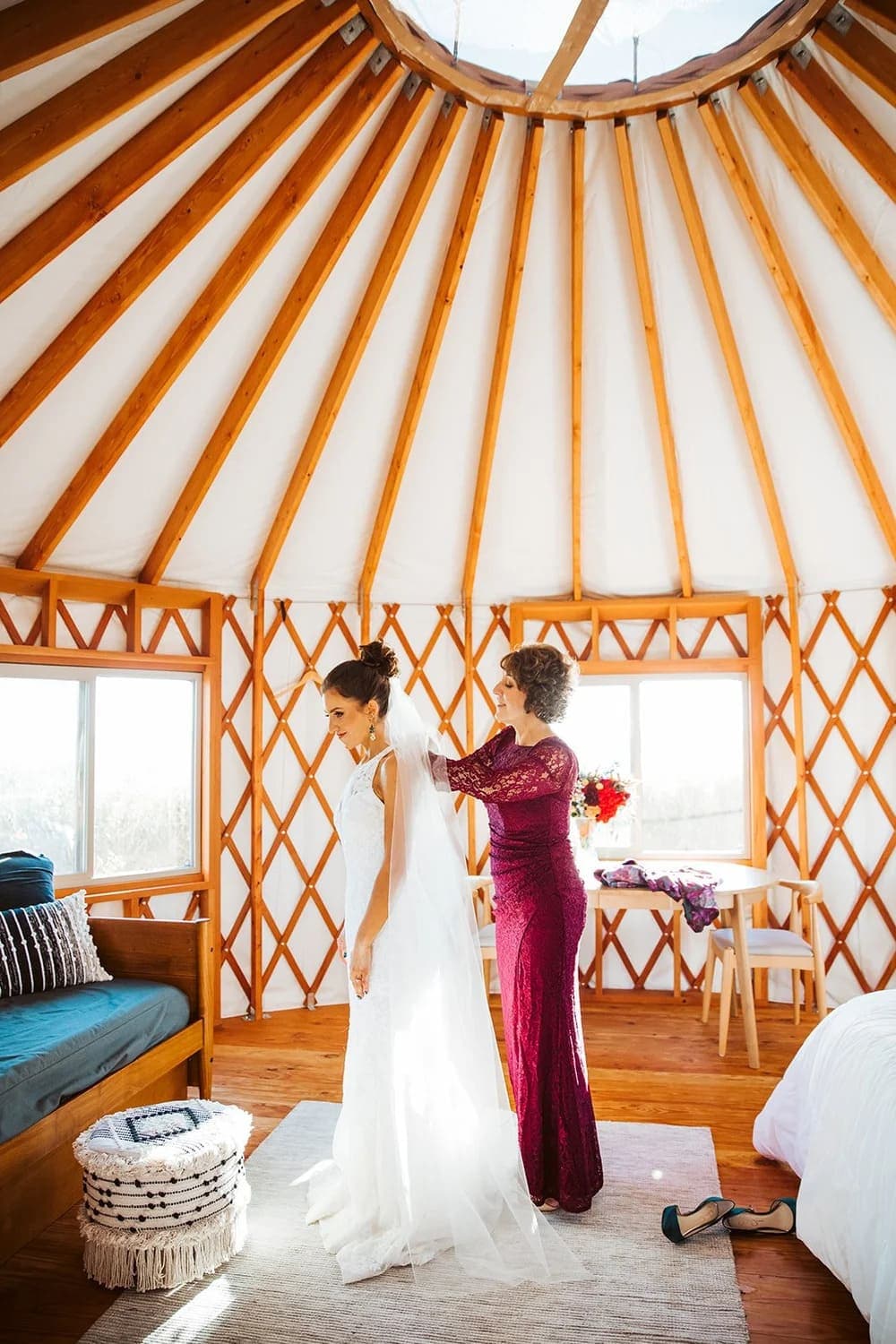 A bride in a white gown is being helped with her veil by a woman in a burgundy dress inside a cozy yurt.