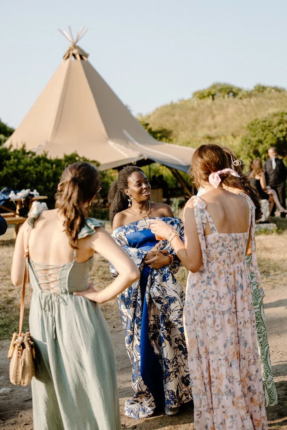 A group of women in elegant dresses chatting outdoors near a tent.