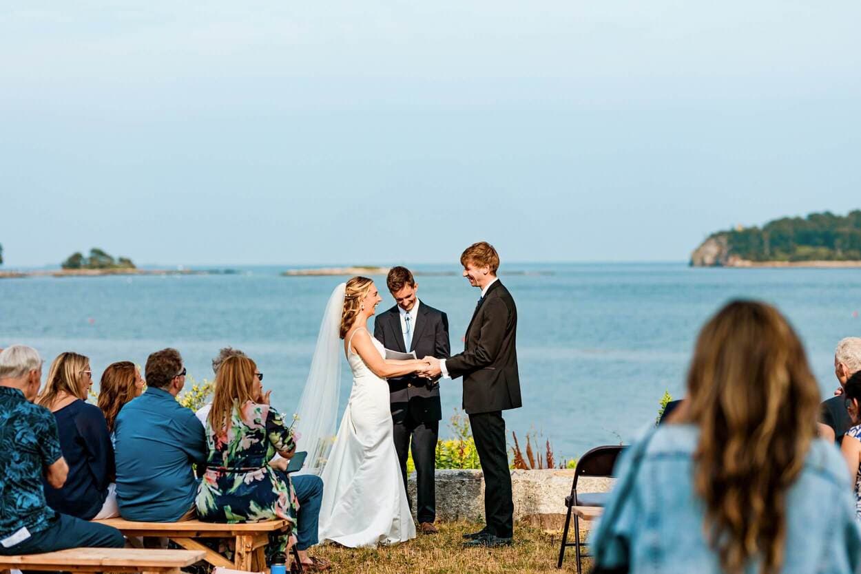 A bride and groom exchange vows during an outdoor wedding ceremony by the water, surrounded by guests.