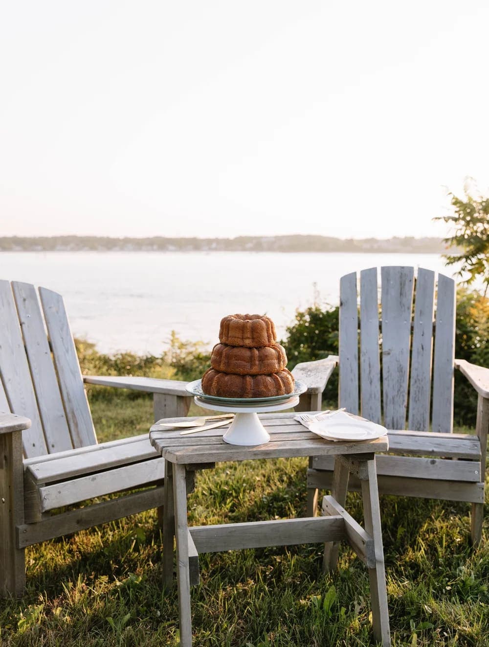 A three-tiered cake is displayed on a table between two wooden chairs, with a scenic view of water in the background.