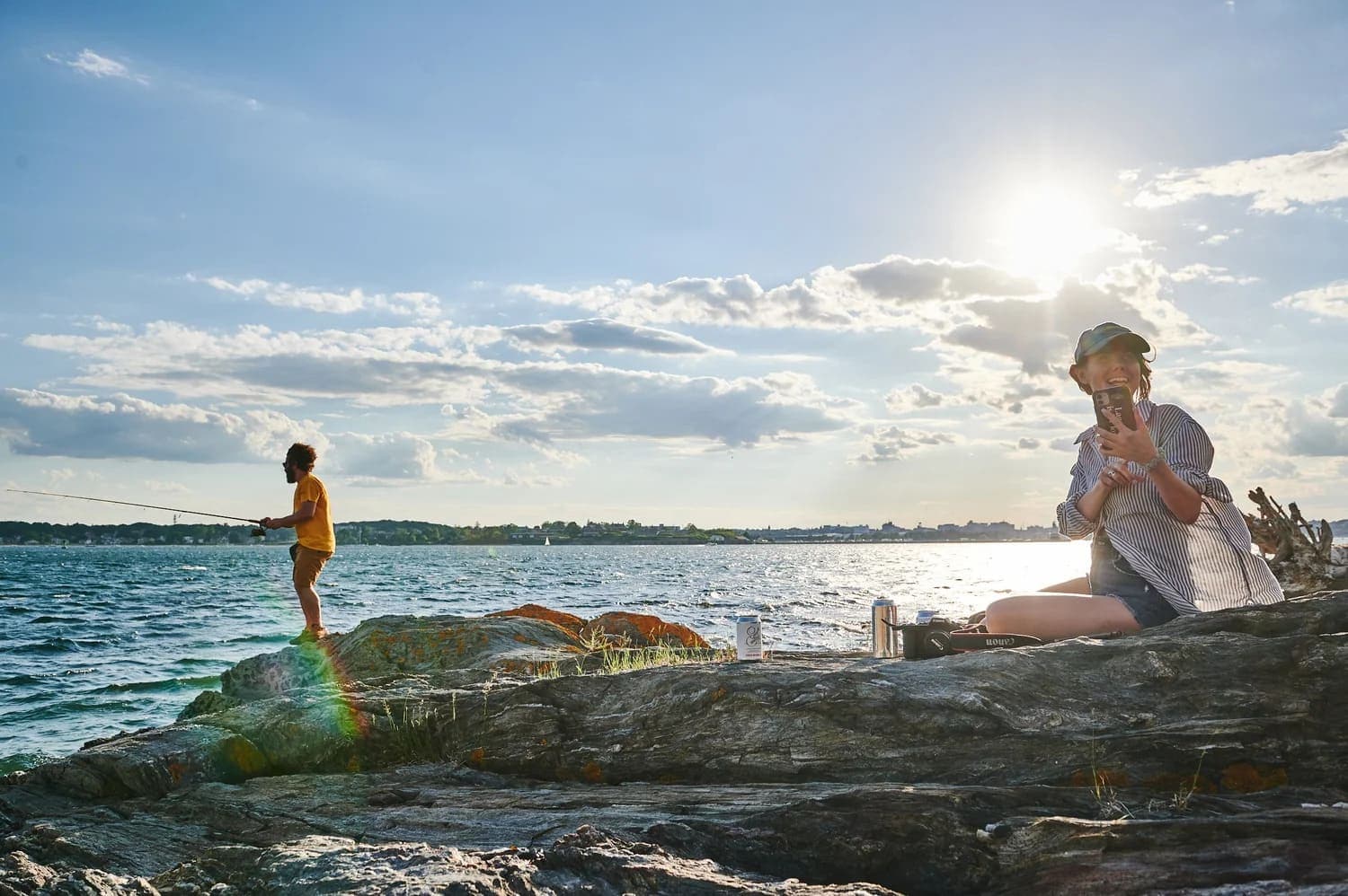 A person sitting on rocks smiles at the camera while another person fishes in the background under a sunny sky.