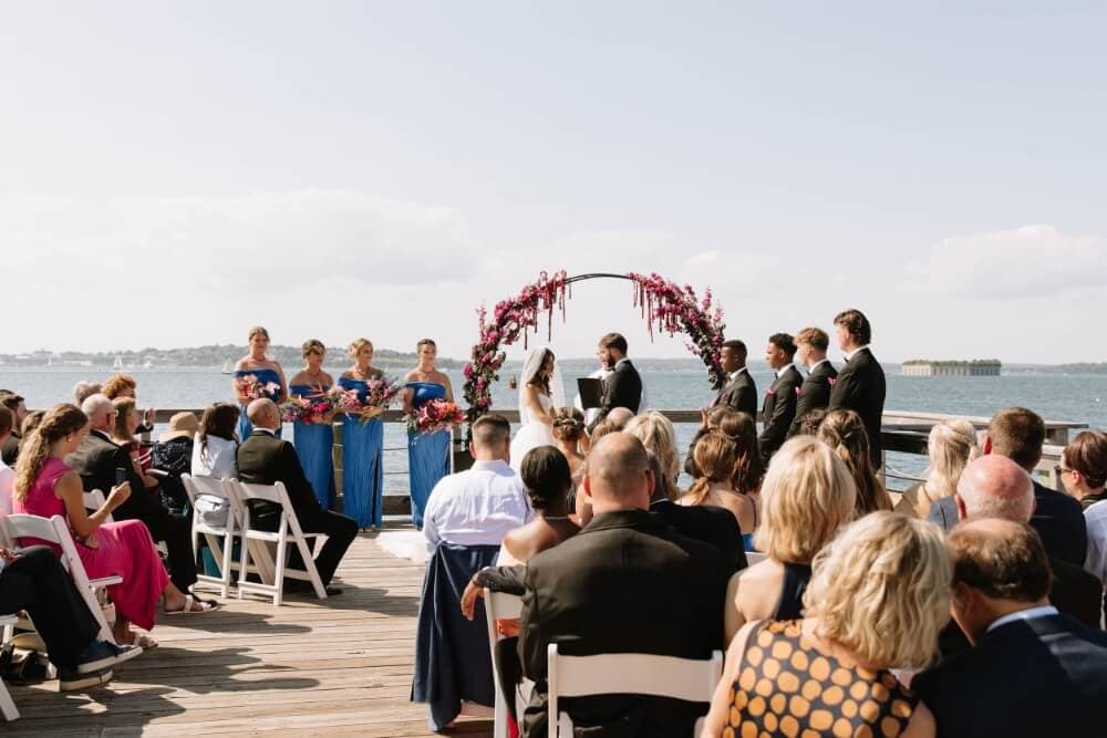A wedding ceremony is taking place outdoors by the water, with a floral arch and guests seated in attendance.