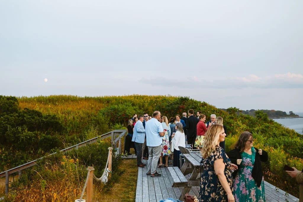 A group of people socializes on a wooden deck overlooking a grassy hillside and the water at dusk.