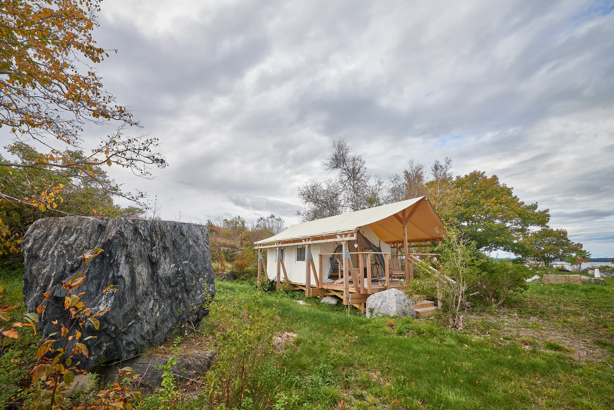 A wooden cabin with a canvas roof beside a large boulder in a grassy landscape under a cloudy sky.