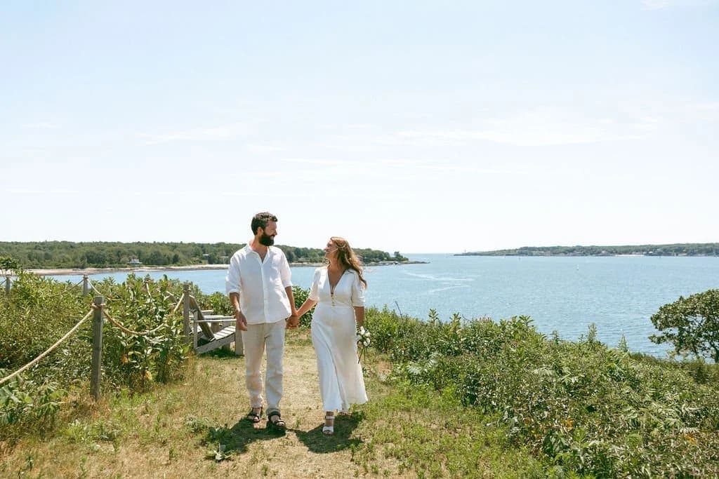 A couple walks hand in hand along a coastal path with a serene water view.