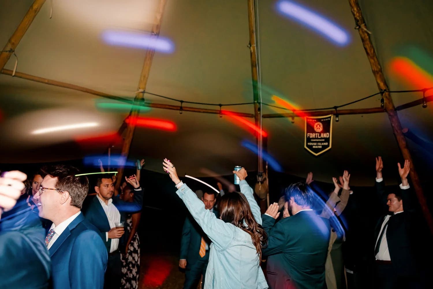A lively group of people dances inside a tent, illuminated by colorful lights.